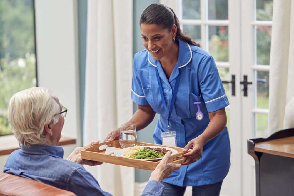 Female Care Worker In Uniform Bringing Meal On Tray To Senior Man Sitting In Lounge