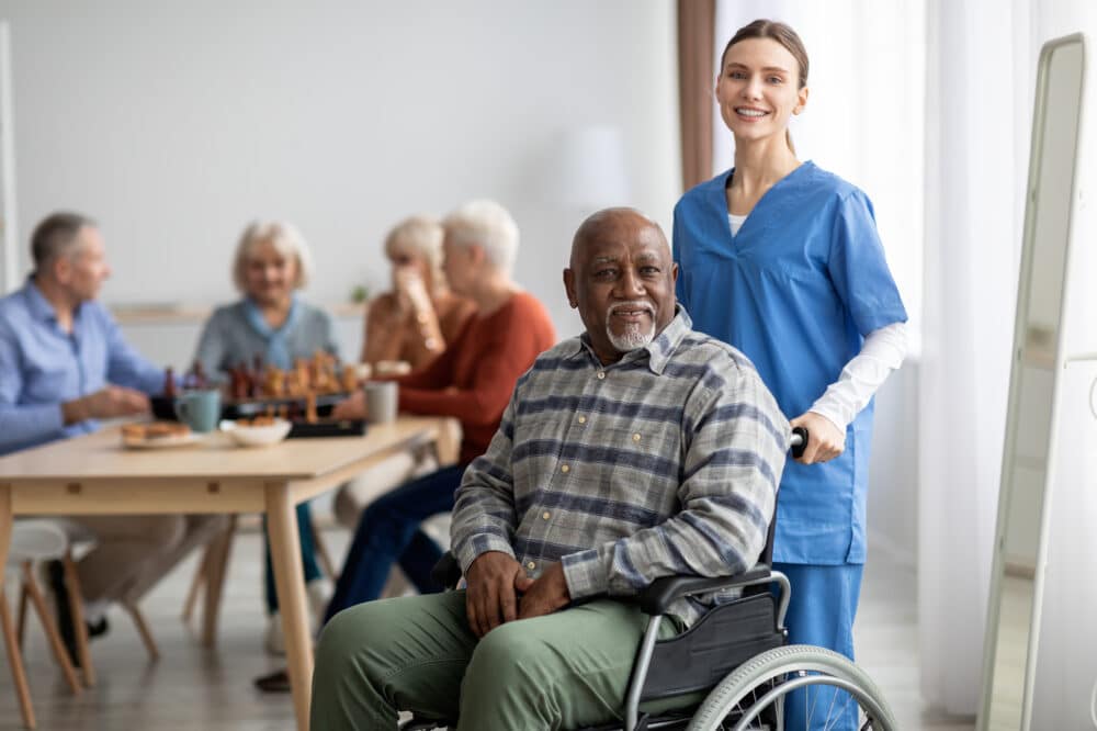 Happy black man older patient on wheelchair with female nurse smiling at camera, group of senior people sitting on couch on background, memory care for elderly people concept