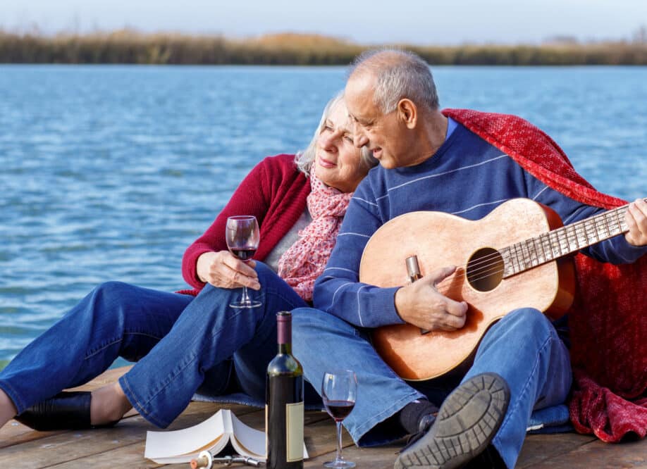 Cottage Living. Happy senior couple enjoying time together playing guitar and drinking wine by the lake wrap around in a red blanket.