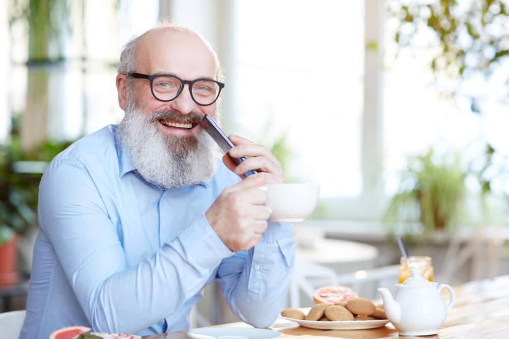 Happy mature man in eyeglasses and blue shirt looking at camera by table with cookies and teapot