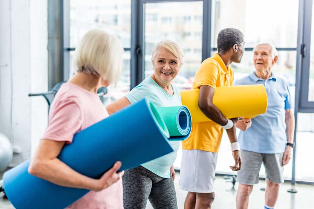 Senior sportswomen holding fitness mats and their male friends standing behind at gym. Independent Living Community.