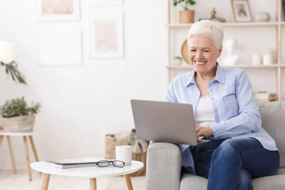 A Happy Elderly Woman With A Laptop At Home, Sitting On The Sofa In The Living Room