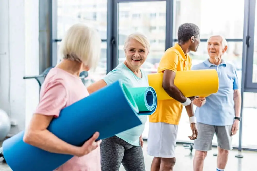 senior sportswomen holding fitness mats and their male friends standing behind at the gym
