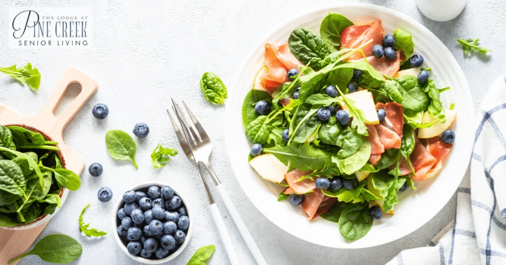 A vibrant salad featuring fresh spinach leaves, thin slices of prosciutto, juicy blueberries, and crisp pear pieces is neatly arranged on a white plate. The scene also includes a small bowl of blueberries and scattered spinach leaves around the plate, highlighting the importance of nutritious foods for memory care.