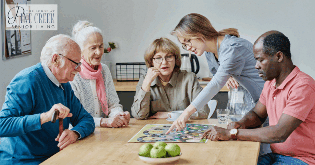 A group of seniors at a memory care community sits around a table playing a board game, while a caregiver explains the rules and provides support. The scene shows engagement, interaction, and a warm, supportive atmosphere.