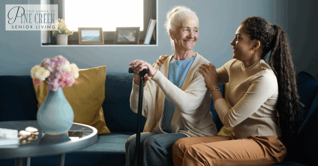 A smiling elderly woman sits on a couch with a cane while her granddaughter lovingly embraces her at a memory care community. The setting is cozy and intimate, reflecting emotional connection and attentive care.