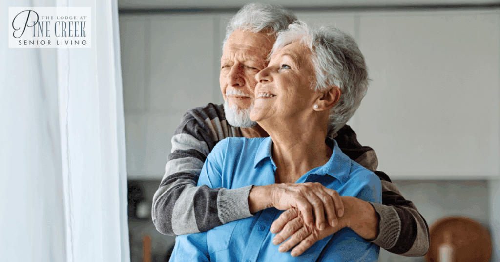 An older couple stands by a bright window as the man gently wraps his arms around the woman from behind, both appearing peaceful and content. This scene reflects the comfort and support often sought in Alzheimer’s care at home.